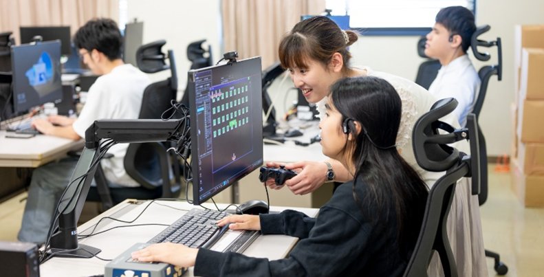 女子学生1名が座ってコンピュータを操作しており、もう1名の女子学生がモニタをのぞき込んでいる。教室の風景。